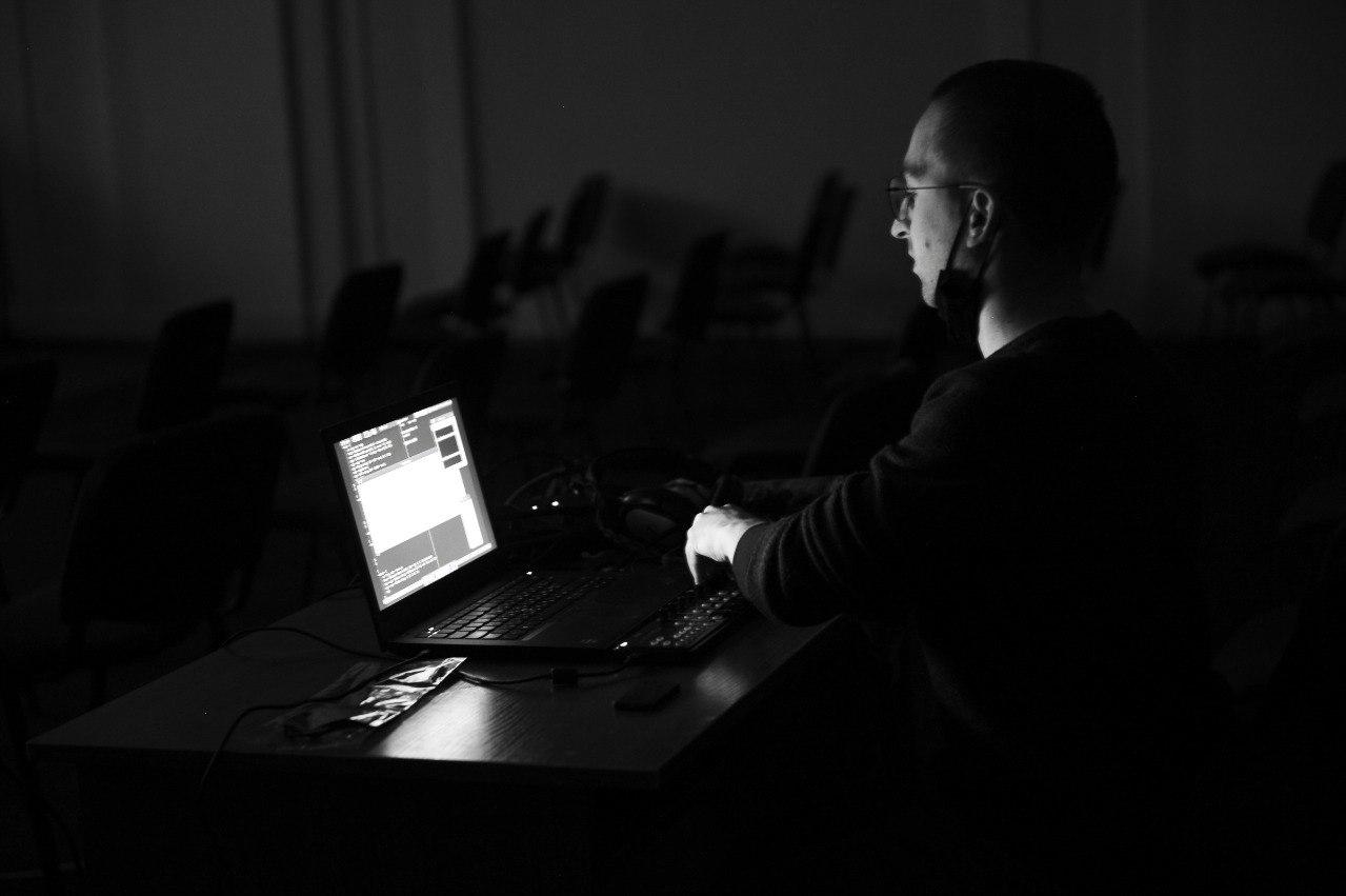 Grayscale photo; person performing sitting in front of a laptop, turning knobs on a midi controller, in a dark concert hall. Empty chairs visible in background. Screen glow light the performer's face.
